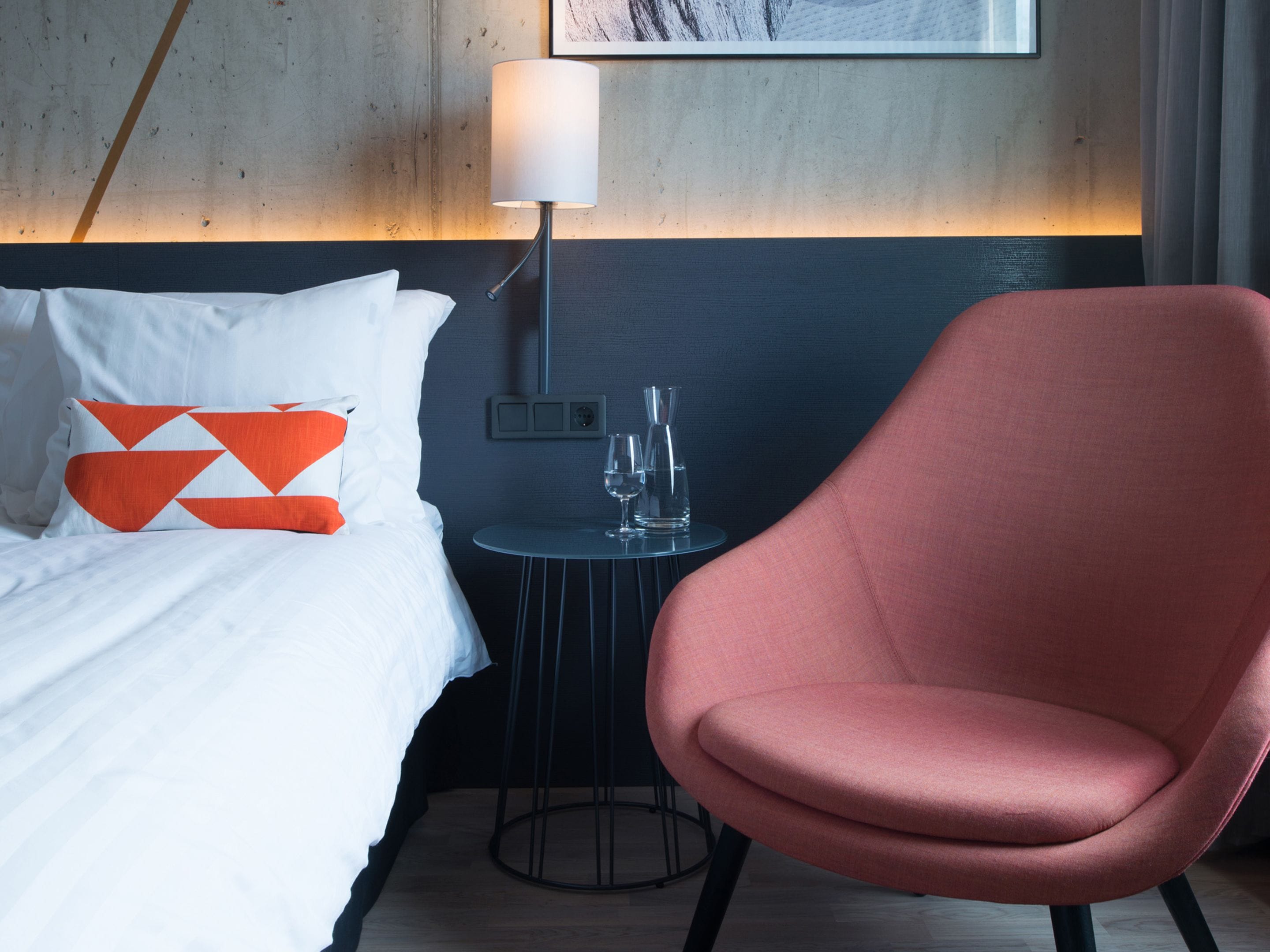 Une chambre d’hôtel avec un lit, un fauteuil rouge, une petite table avec un verre et une lampe.