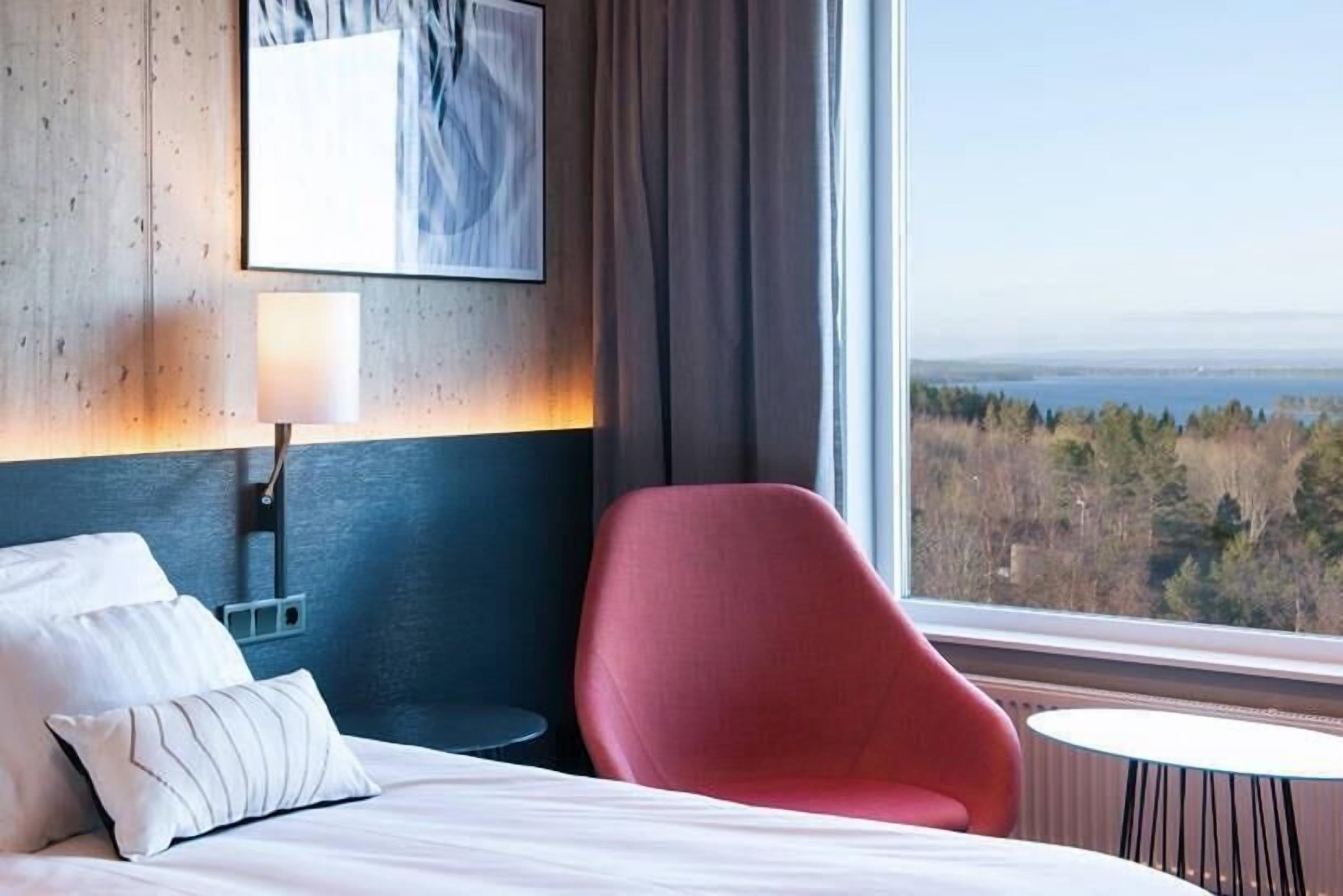 A hotel room with a bed, a red chair, a small table, a lamp, and a window with a view of trees and a body of water.