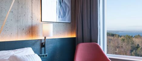 A hotel room with a bed, a red chair, a small table, a lamp, and a painting on the wall.