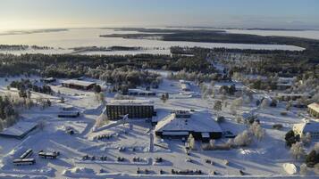 A snow-covered landscape with a hotel and several buildings, surrounded by trees and a body of water in the distance.