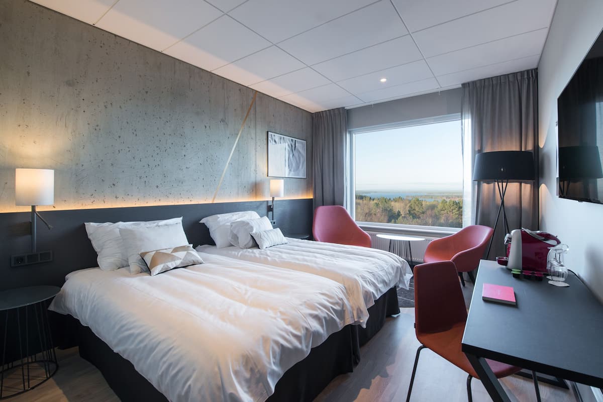 A modern hotel room with a large bed, two red chairs, a small table, and a view of the outdoors.