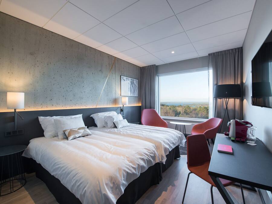 Een moderne hotelkamer met een groot bed, twee rode stoelen, een tafeltje en uitzicht op het groen.