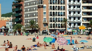 Una playa con gente tomando el sol y un edificio grande con varios balcones al fondo.