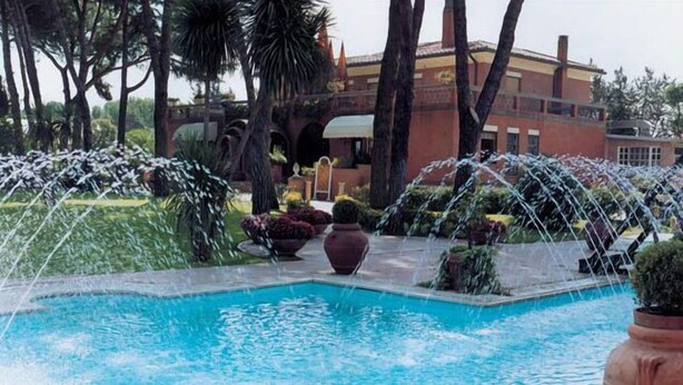 A swimming pool with water jets, surrounded by palm trees and a building with arches in the background. - Hotel Il Quadrifoglio (Rome)
