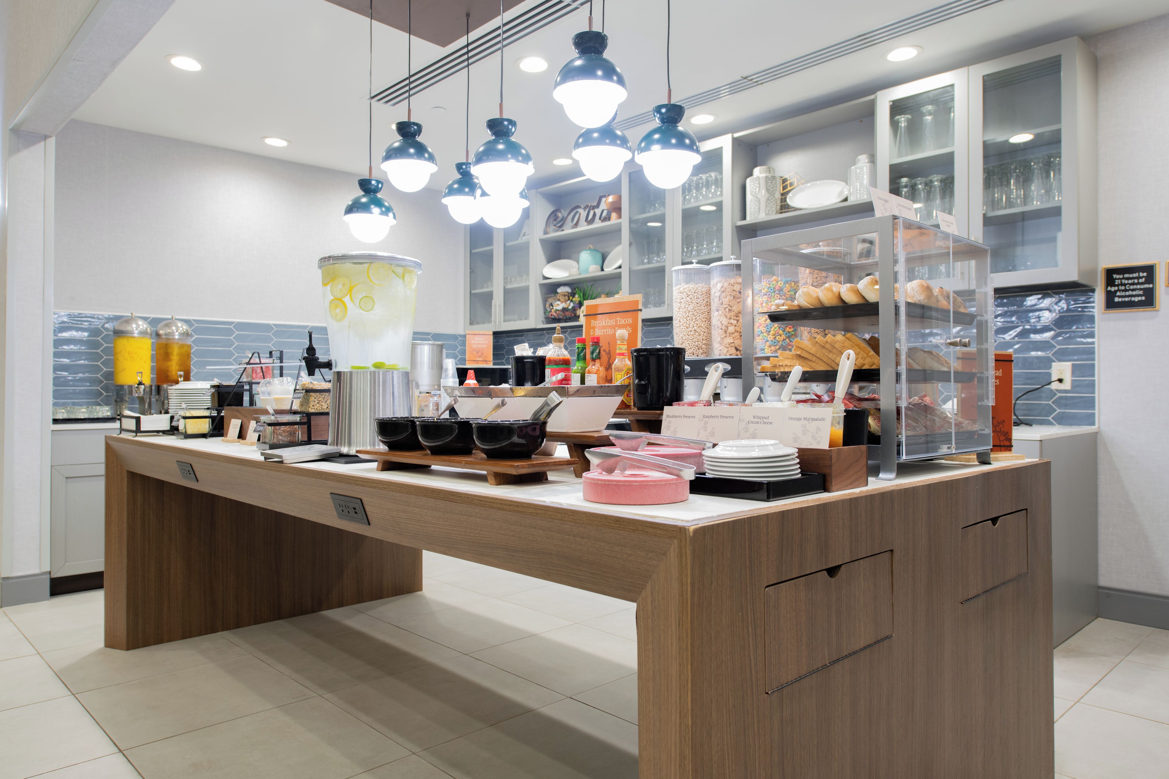 Una barra de desayuno con diversas opciones de comida y bebidas, incluyendo una jarra grande de té helado, tazones de cereales y una selección de pasteles.