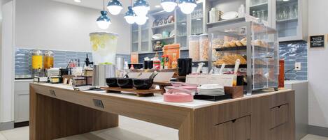Una barra de desayuno con diversas opciones de comida y bebidas, incluyendo una jarra grande de té helado, tazones de cereales y una selección de pasteles.