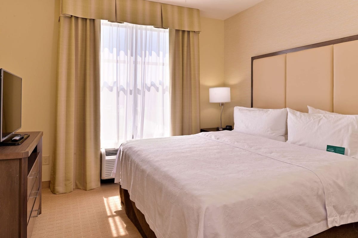 A hotel room with a bed, a television on a desk, a lamp, and a window with curtains.