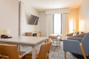 A hotel room with a dining table, two chairs, a sofa, a TV, and a window with curtains. - Homewood Suites Houma, La (Houma)