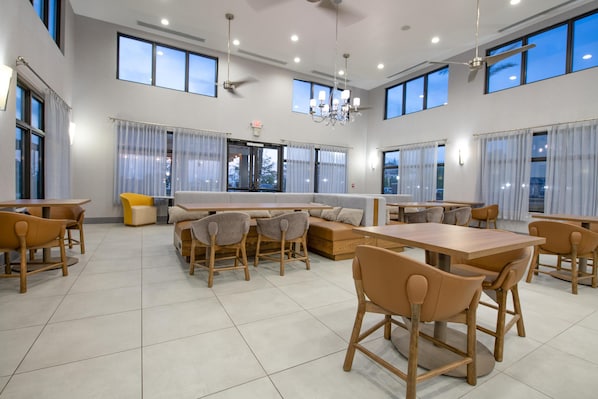 A spacious dining area with multiple tables and chairs, large windows, and a chandelier.