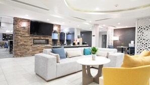 A modern hotel lobby with a stone accent wall, a flat-screen TV, a sofa with cushions, a round coffee table, and a yellow armchair. - Homewood Suites Houma, La (Houma)