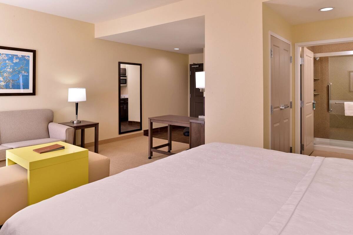 A hotel room with a bed, a desk, a chair, a sofa, a mirror, and a shower.