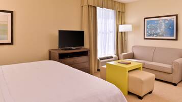 A hotel room with a bed, a desk with a TV, a sofa, and a window with curtains.