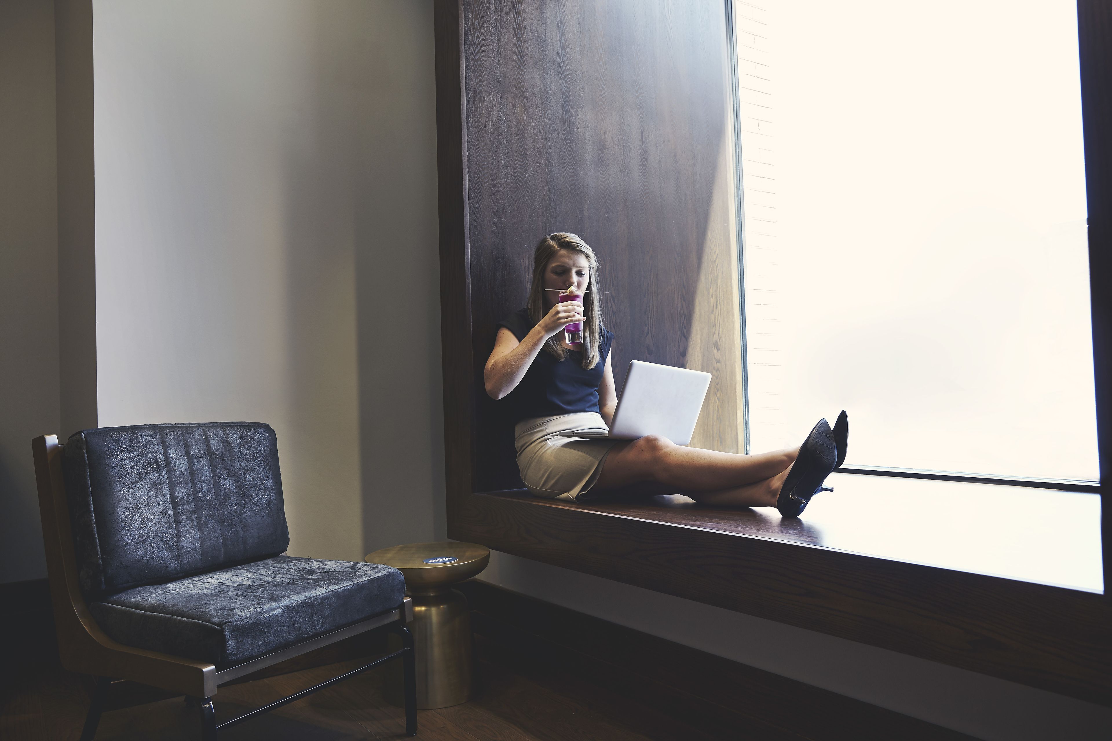 Una persona sentada en el alféizar de una ventana, con una computadora portátil, una silla y una mesita redonda cerca.