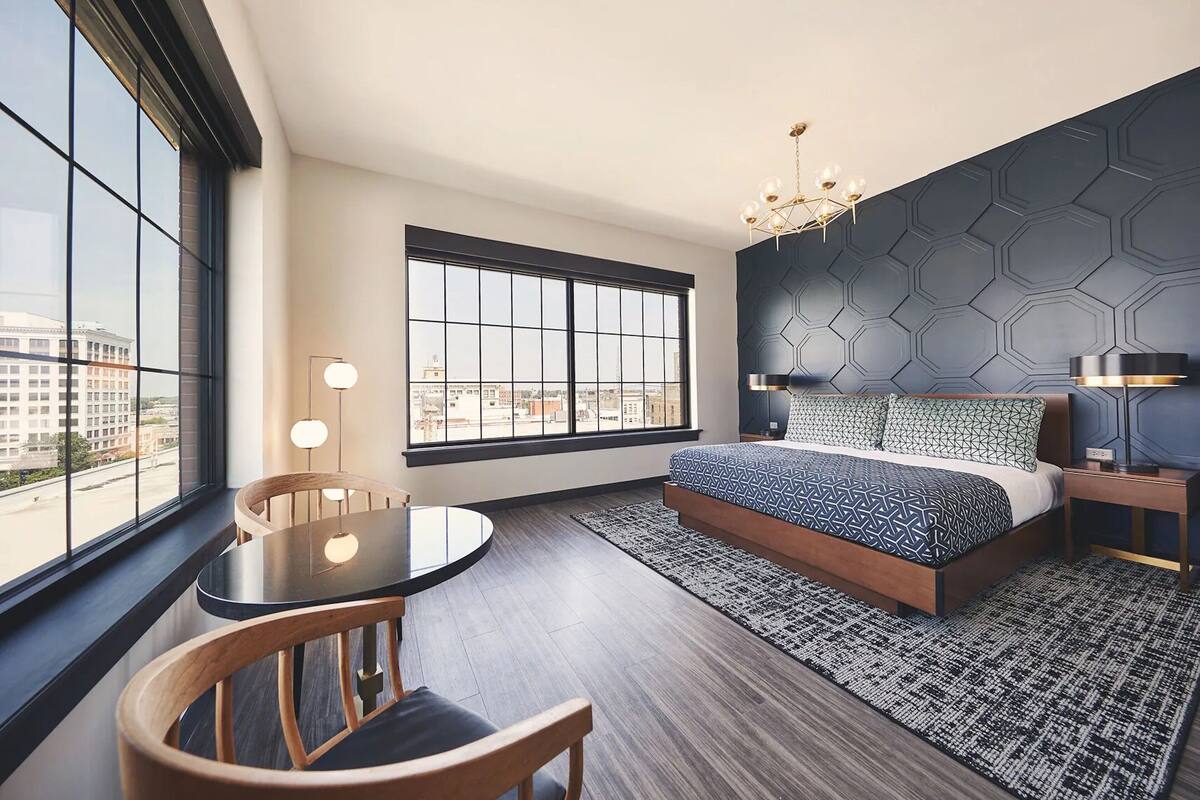 Ett modernt hotellrum med en stor säng, ett runt bord och utsikt över staden genom stora fönster.