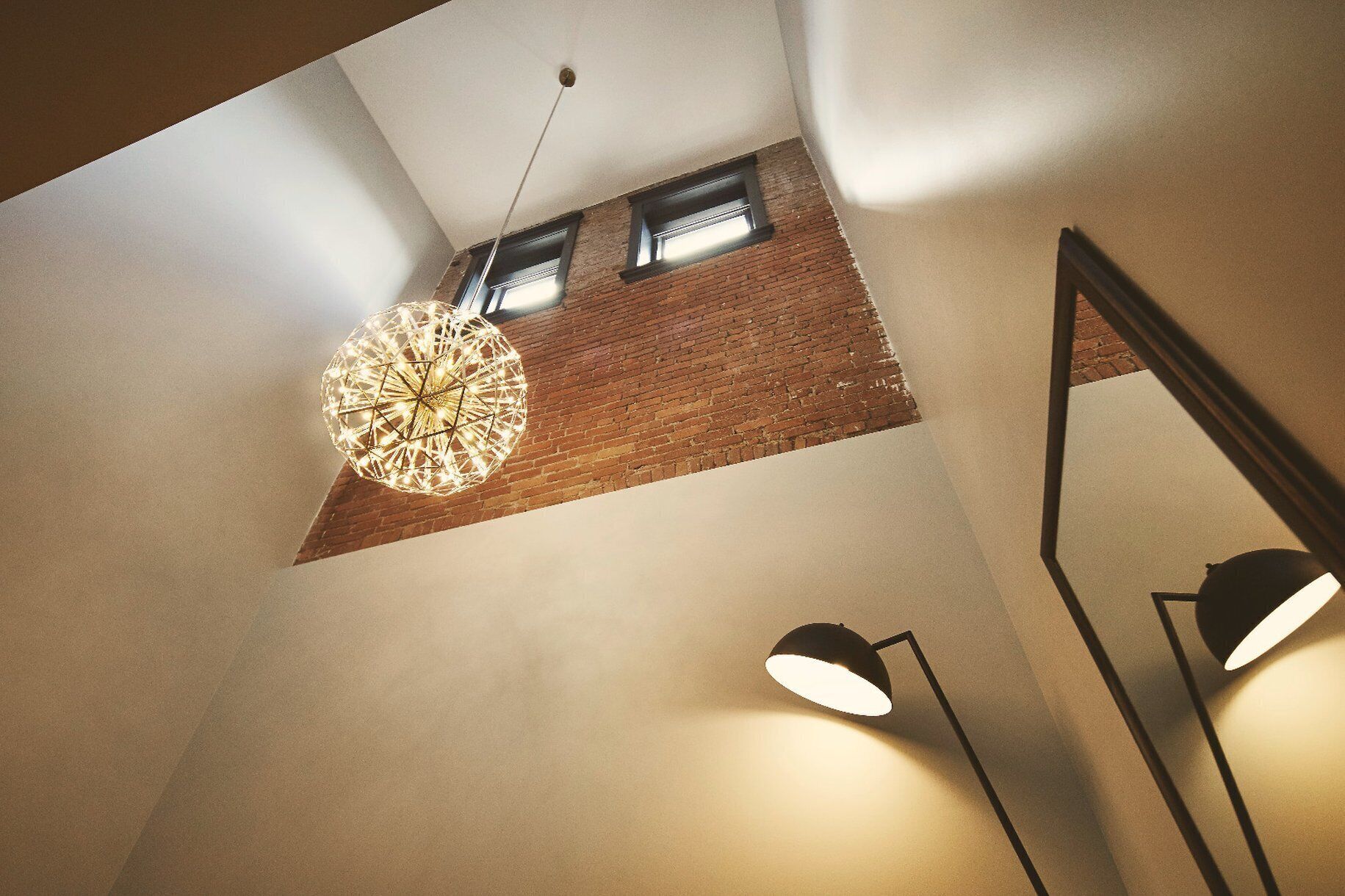 A modern interior with a geometric light fixture, a brick wall, and a large mirror reflecting the room.