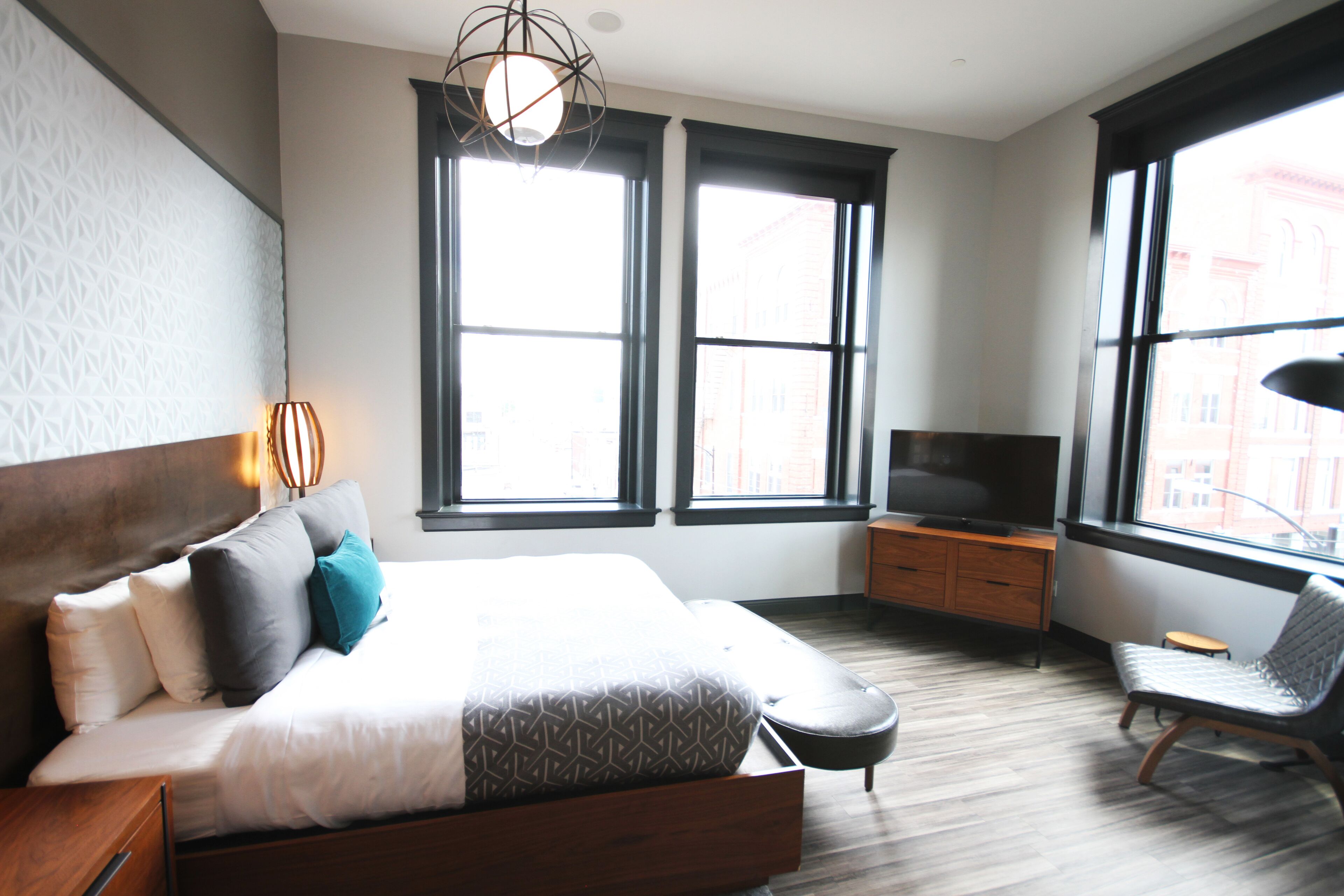 A modern hotel room with a large bed, a TV, a chair, and two windows offering a city view.