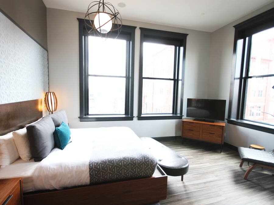 A modern hotel room with a large bed, a TV, a chair, and two windows offering a city view.