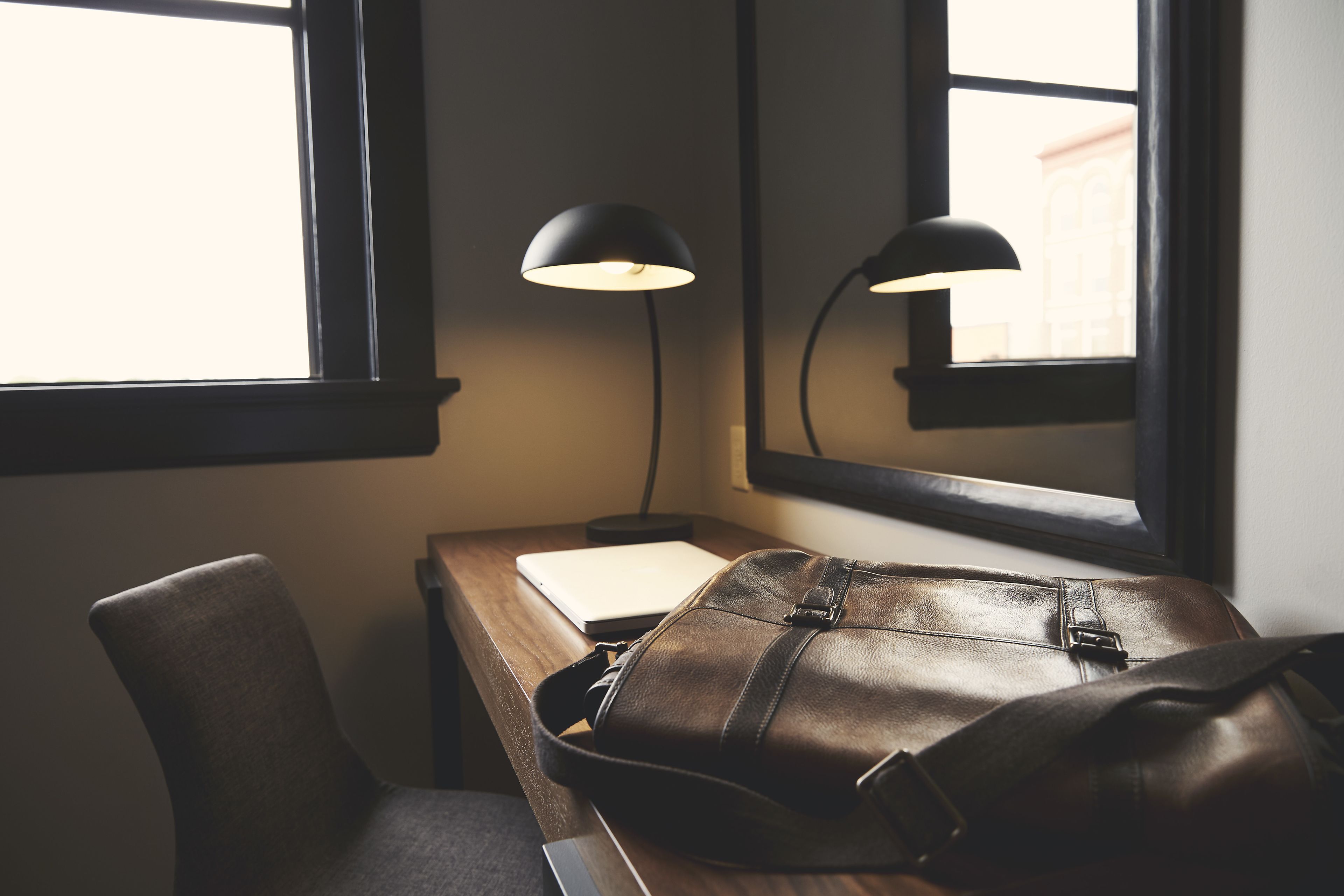 A desk with a leather bag, a notebook, and two desk lamps by a window.