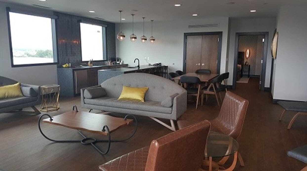 A modern living room with a sofa, coffee table, and chairs. There is a dining area with a table and chairs. A kitchen is visible in the background.