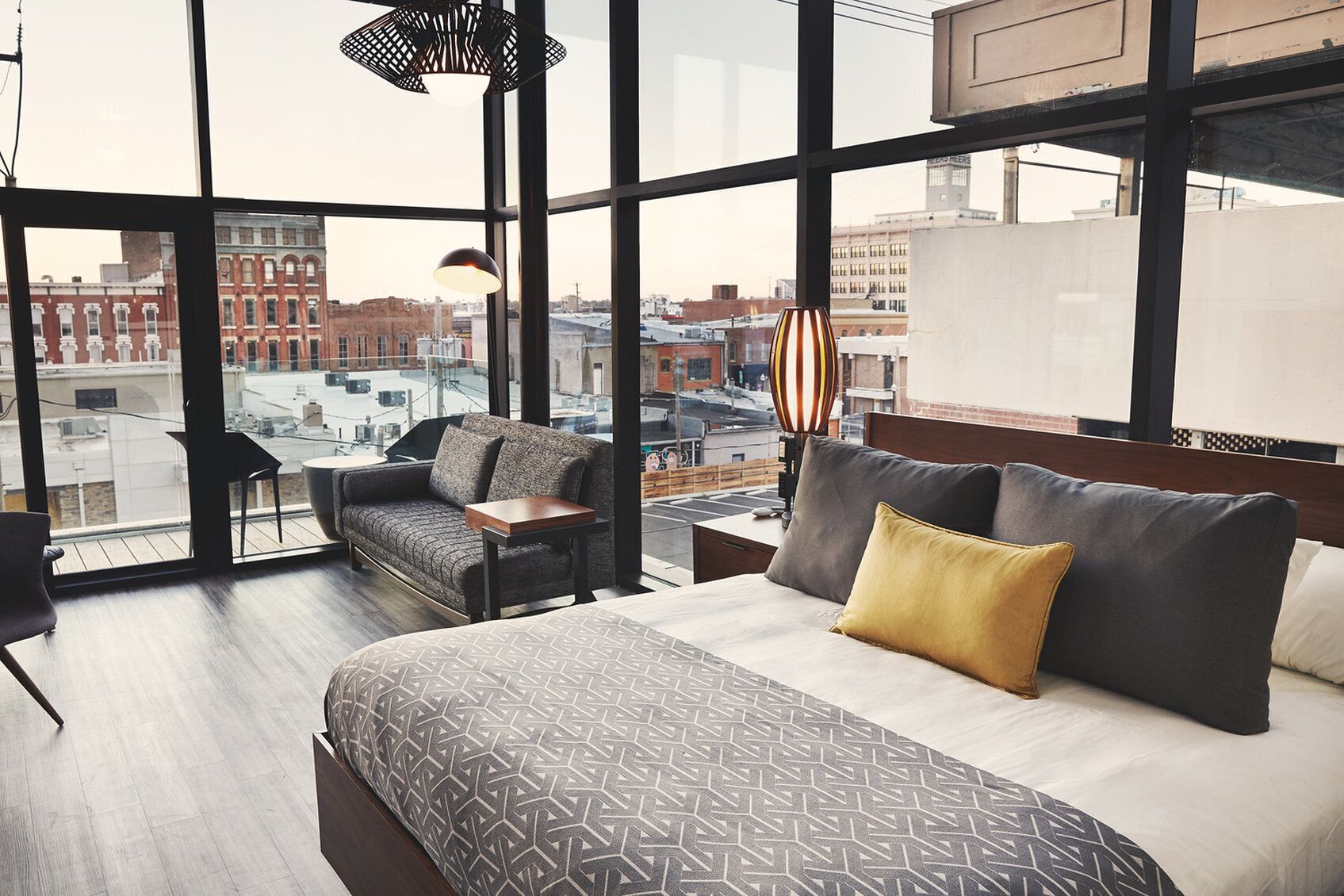 A modern hotel room with a large bed, a sofa, and a small table. The room has large windows offering a city view.