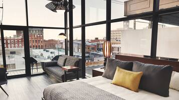A modern hotel room with a large bed, a sofa, and a small table. The room has large windows offering a city view.