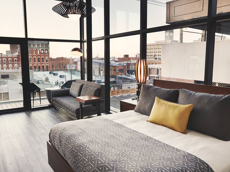 A modern hotel room with a large bed, a sofa, and a small table. The room has large windows offering a city view.