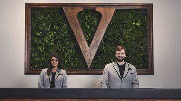Two individuals standing in front of a reception desk with a large V-shaped metal artwork and a green leafy background.