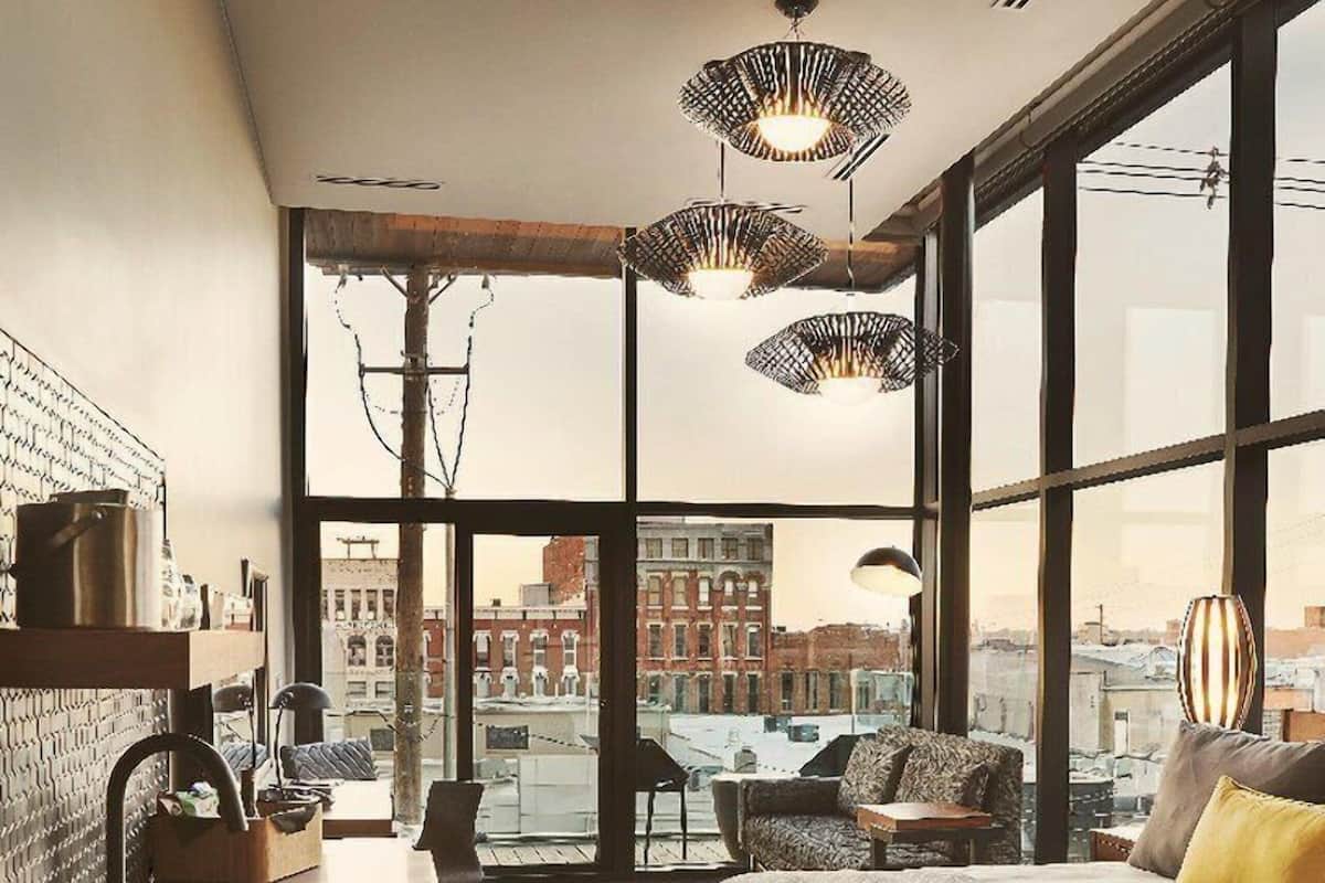 Ett modernt hotellrum med en stor säng, ett skrivbord med handfat och utsikt över stadsbilden genom stora fönster.