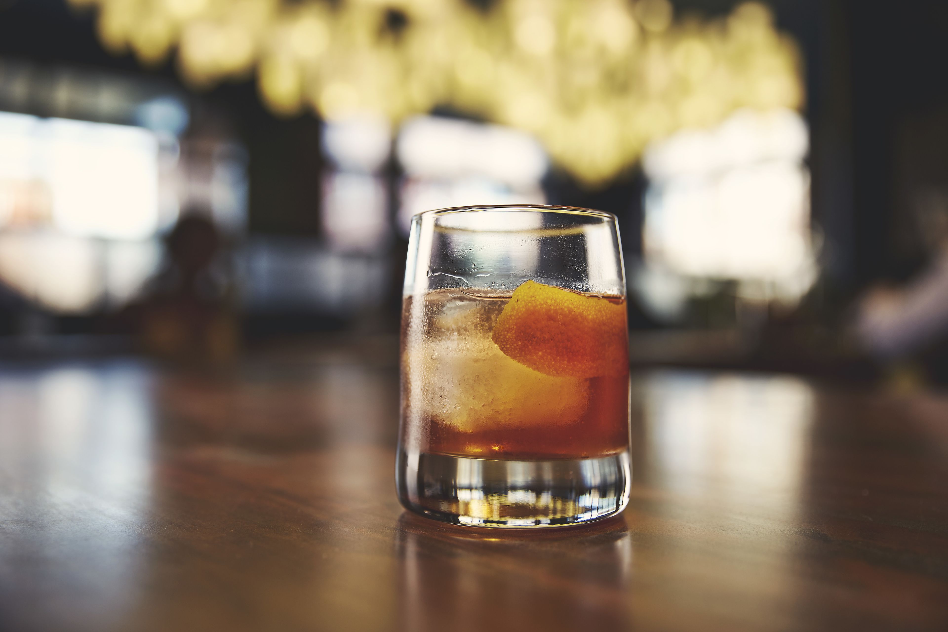 A glass of amber-colored beverage with ice and a slice of orange, placed on a wooden surface.