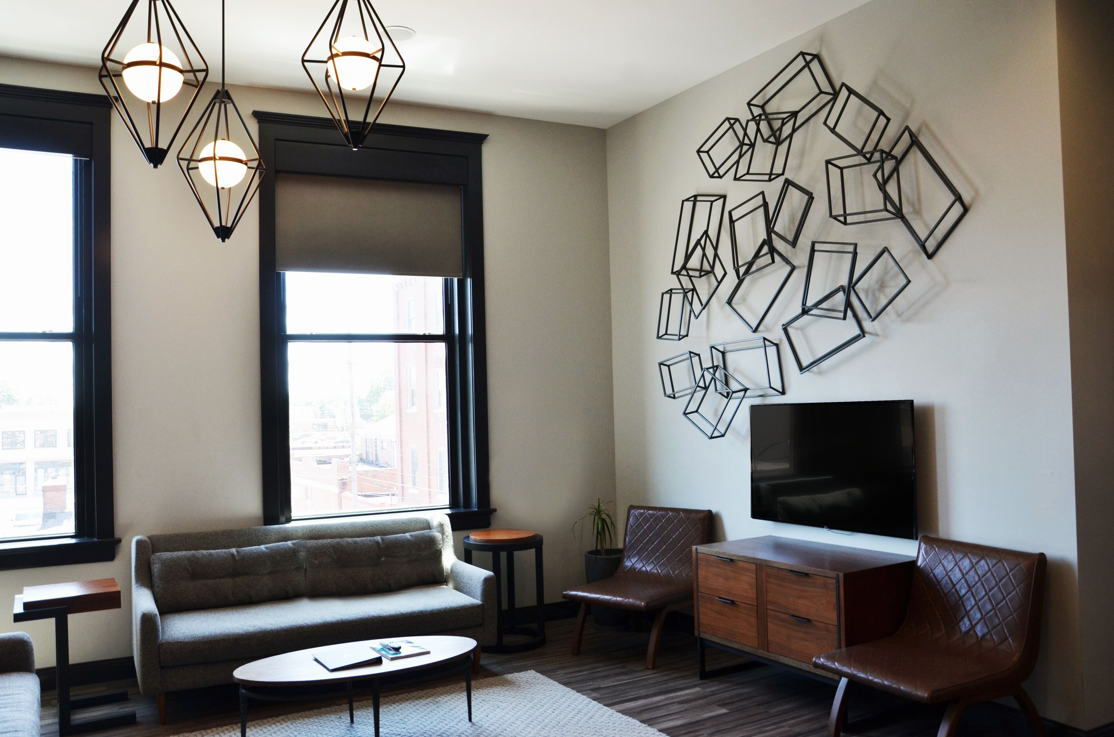A modern living room with a sofa, coffee table, and TV. There are large windows, a potted plant, and a wall with geometric art.