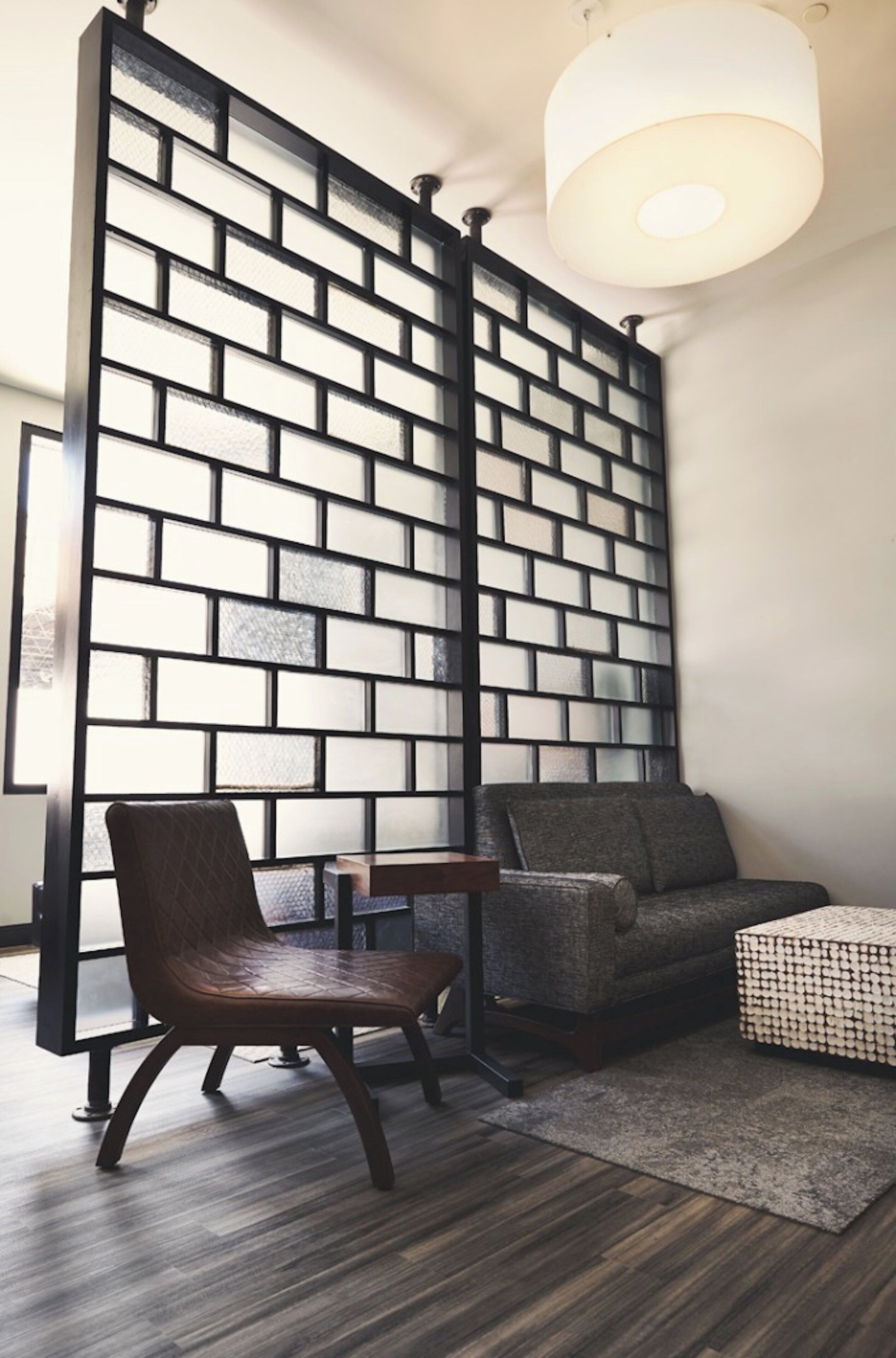 A room with a brick-patterned partition, a wooden chair, a sofa, and a woven ottoman.