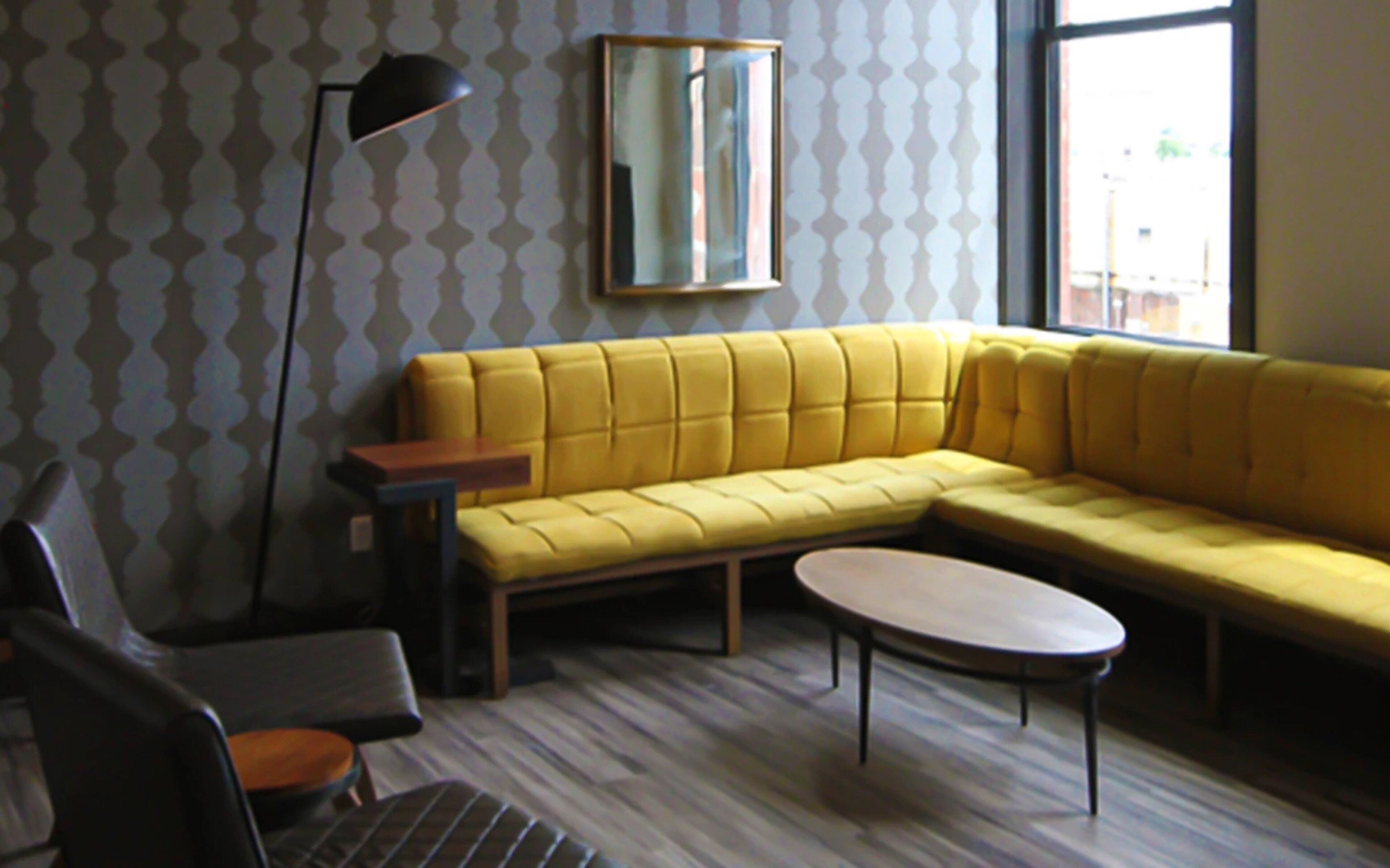 A yellow sectional sofa, a small round table, a floor lamp, and a chair in a room with patterned wallpaper and a window.