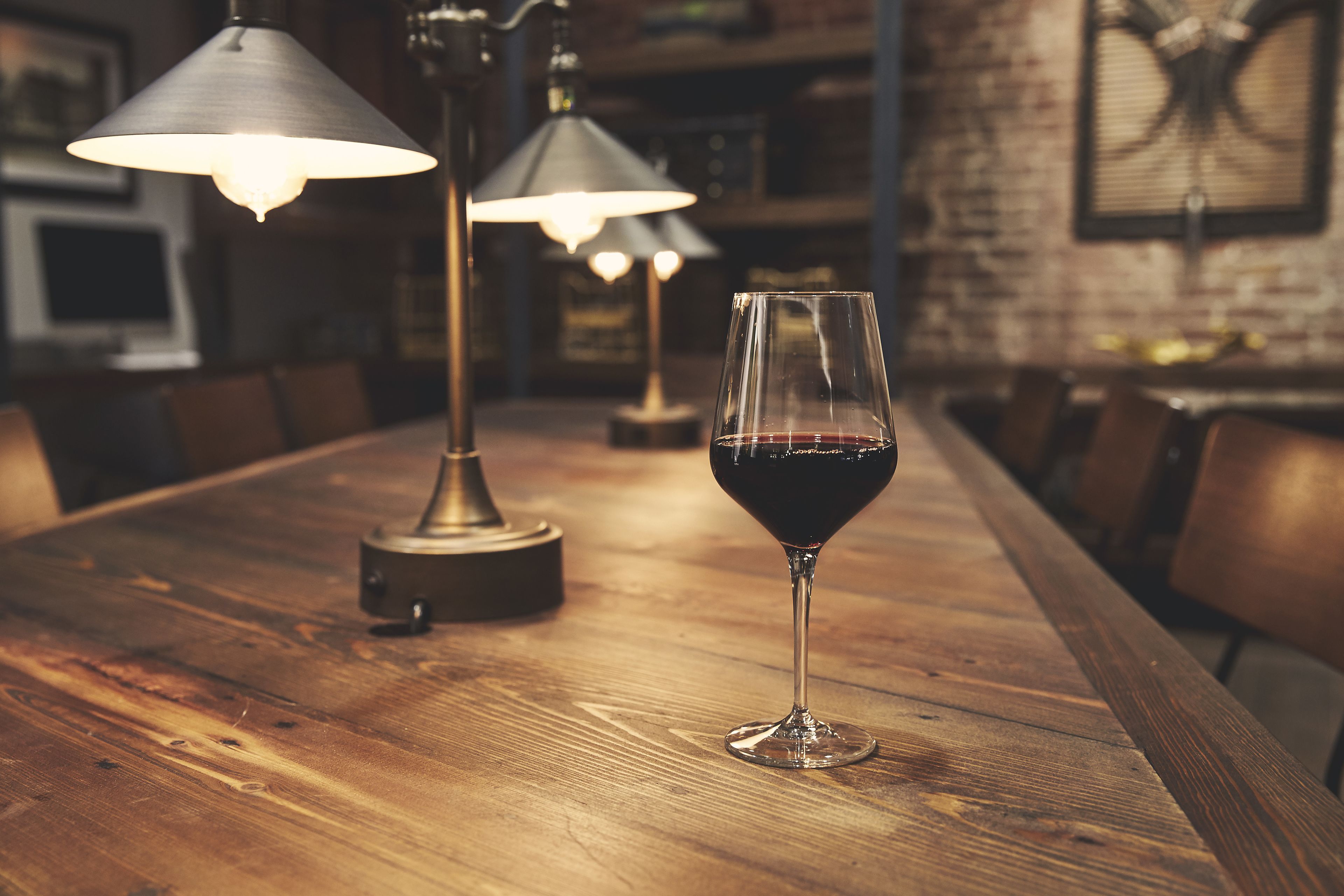 A wooden bar table with a glass of red wine and three lamps in the background.