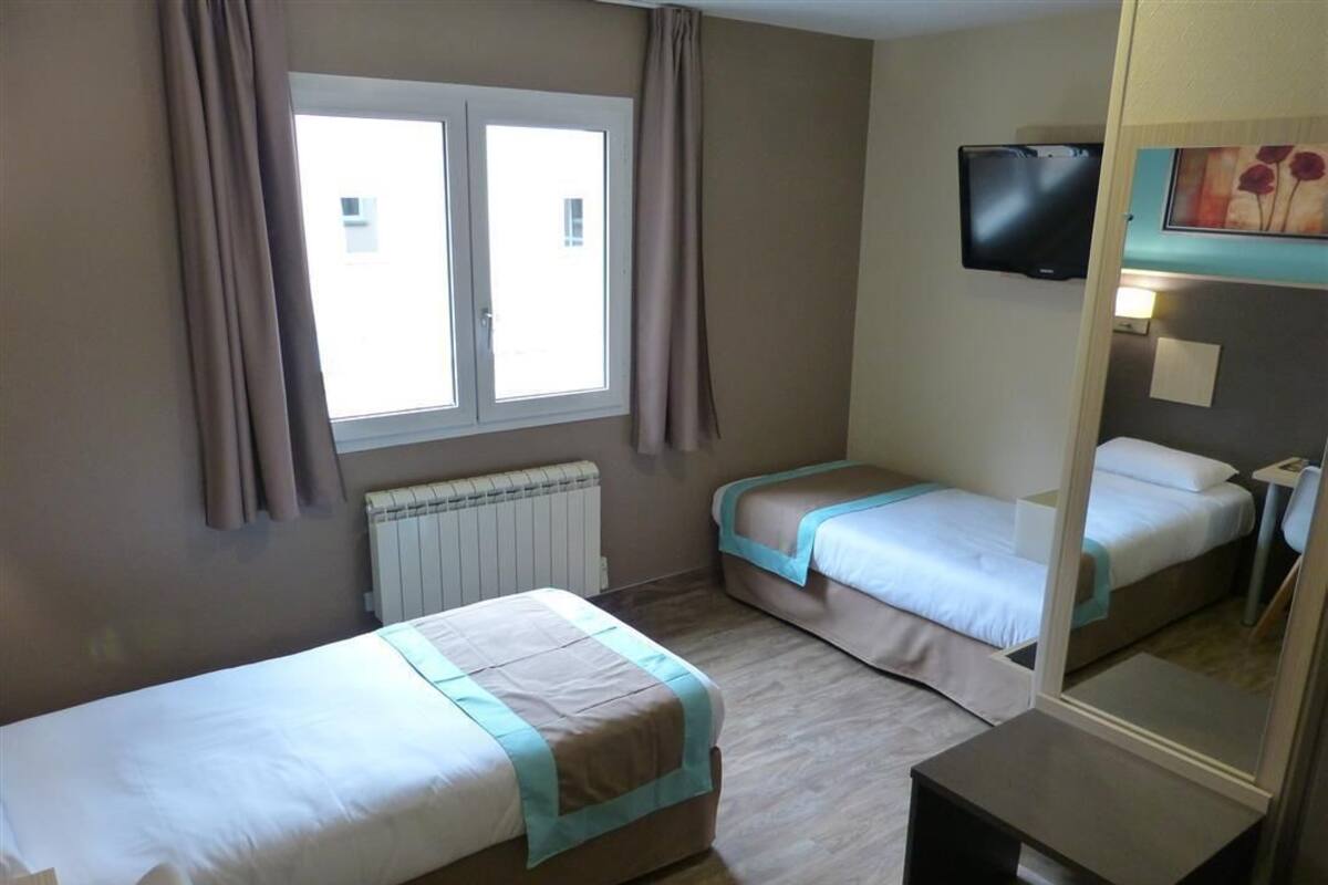 A hotel room with two single beds, a window with curtains, a radiator, a flat-screen TV mounted on the wall, and a small table.