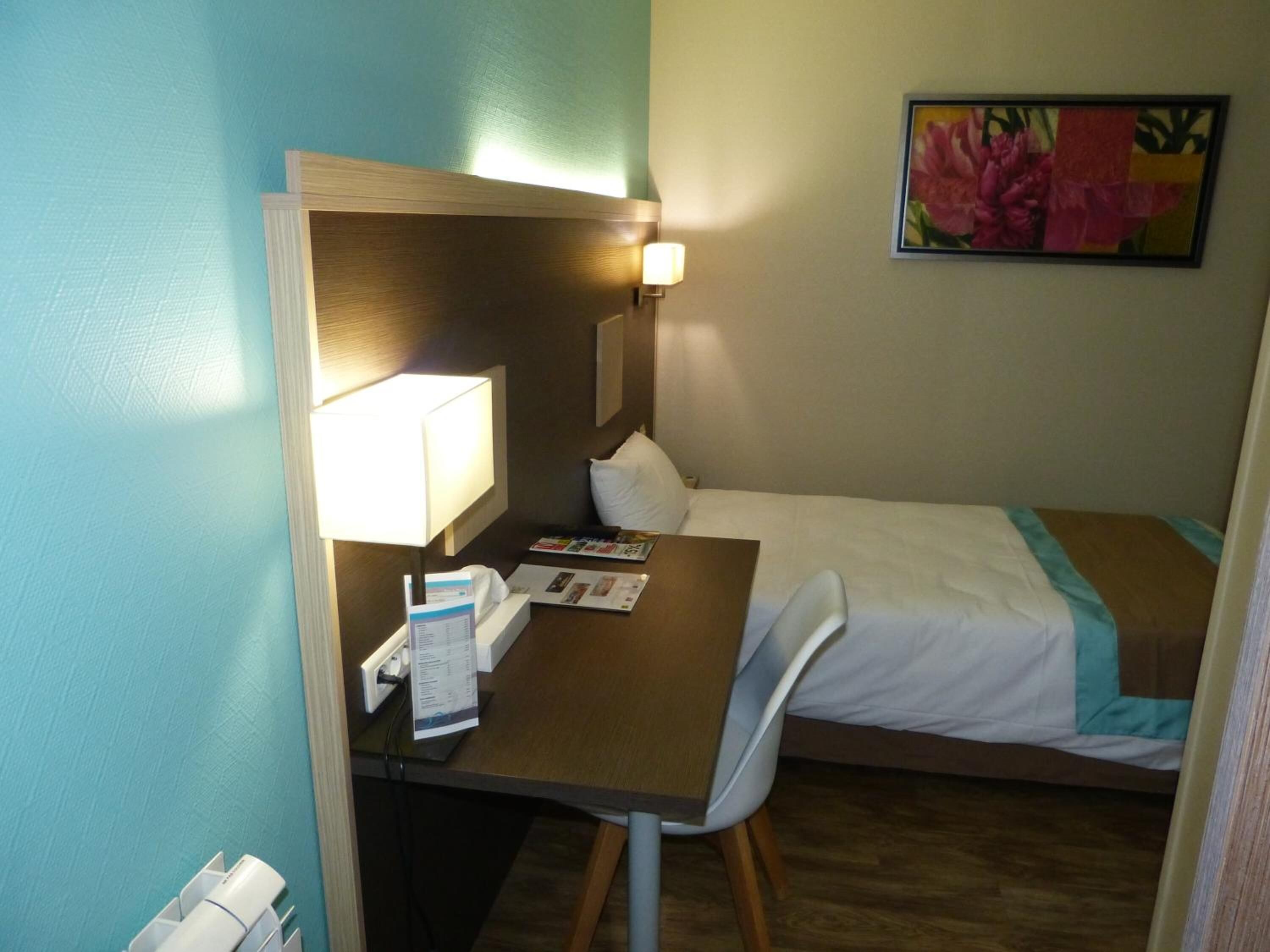 A hotel room with a bed, a desk with a lamp, a chair, and a painting on the wall.