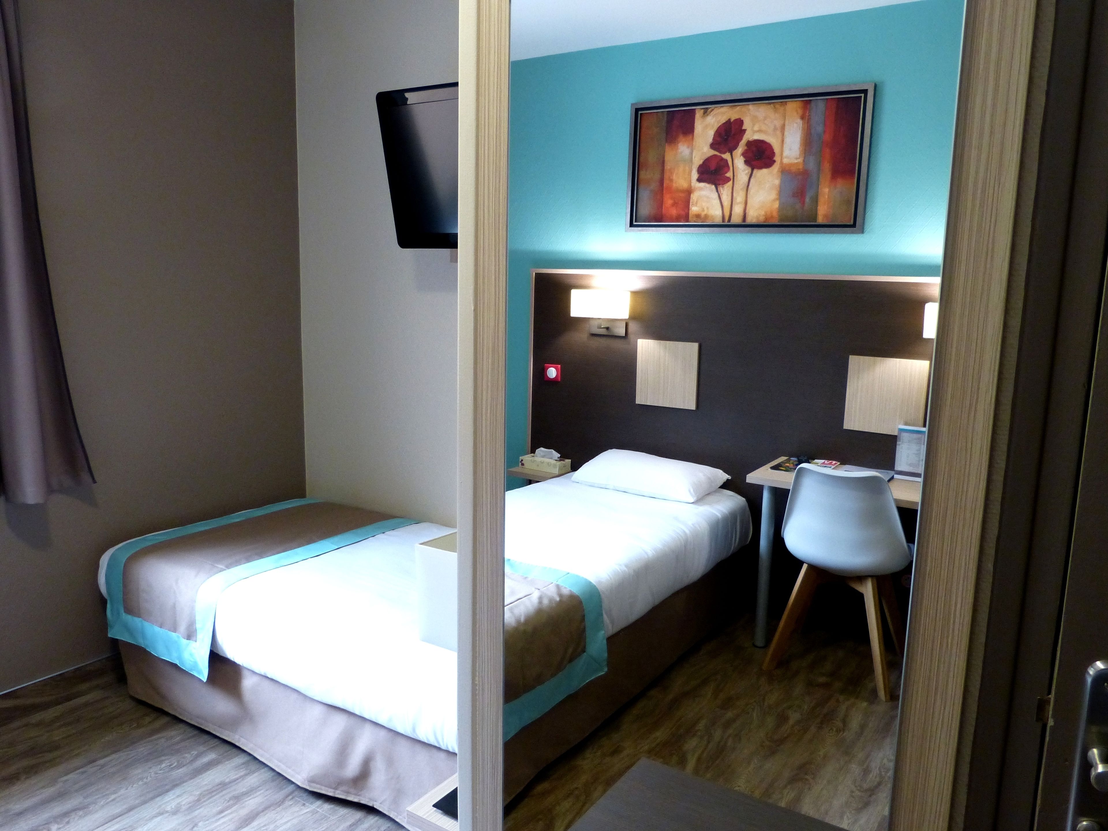 A hotel room with a single bed, a desk, a chair, a TV mounted on the wall, and a painting above the desk.