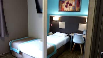 A hotel room with a single bed, a desk, a chair, a TV mounted on the wall, and a painting above the desk.