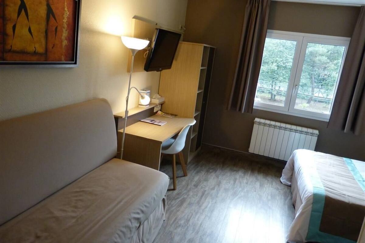A hotel room with a bed, a desk with a chair, a TV, and a window with curtains.