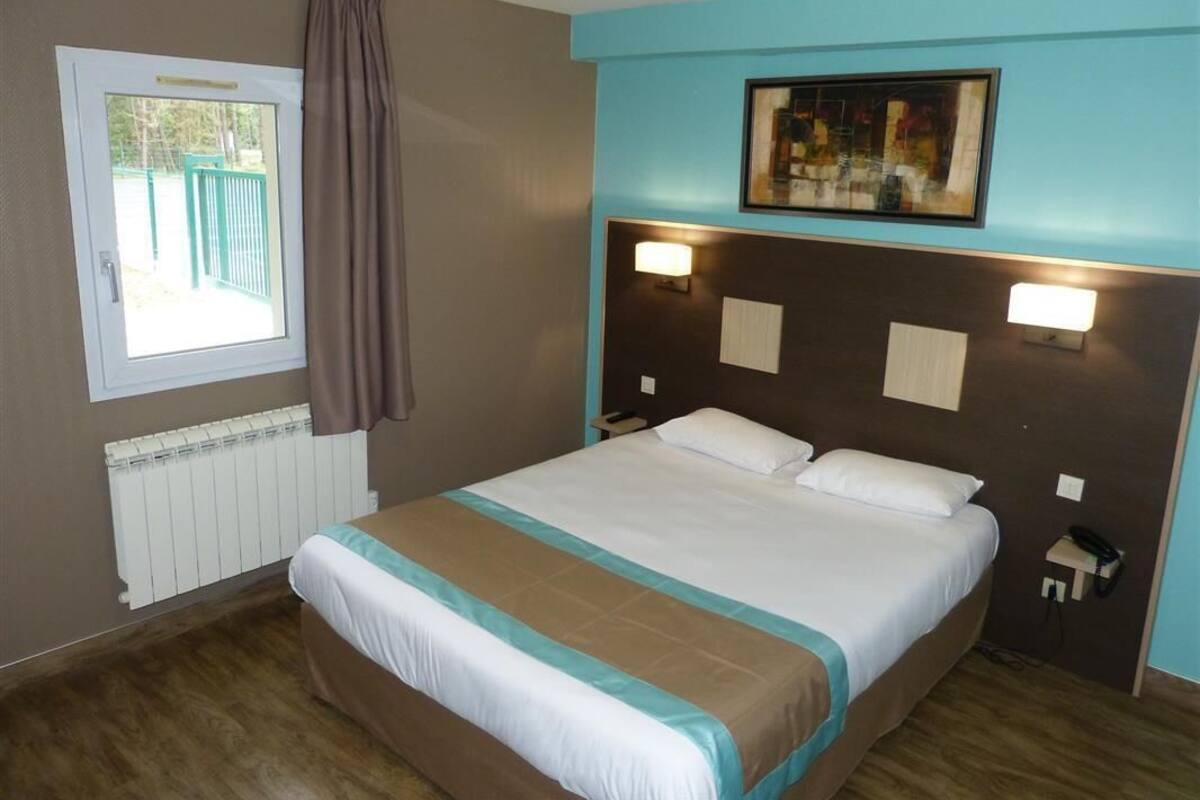 A hotel room with a bed, a window with curtains, a radiator, and a painting on the wall.
