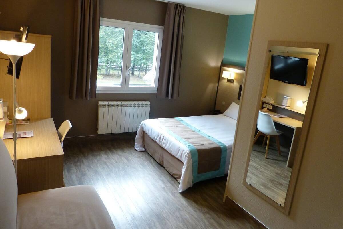 A hotel room with a bed, desk, chair, lamp, window, and TV.