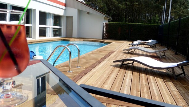 A wooden deck with a swimming pool, lounge chairs, and a glass of drink. - Hôtel Arbor (Mulsanne)