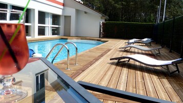 A wooden deck with a swimming pool, lounge chairs, and a glass of drink.