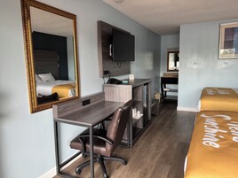 Quarto de hotel com cama, escrivaninha com cadeira, espelho, televisão e mesa de cabeceira.