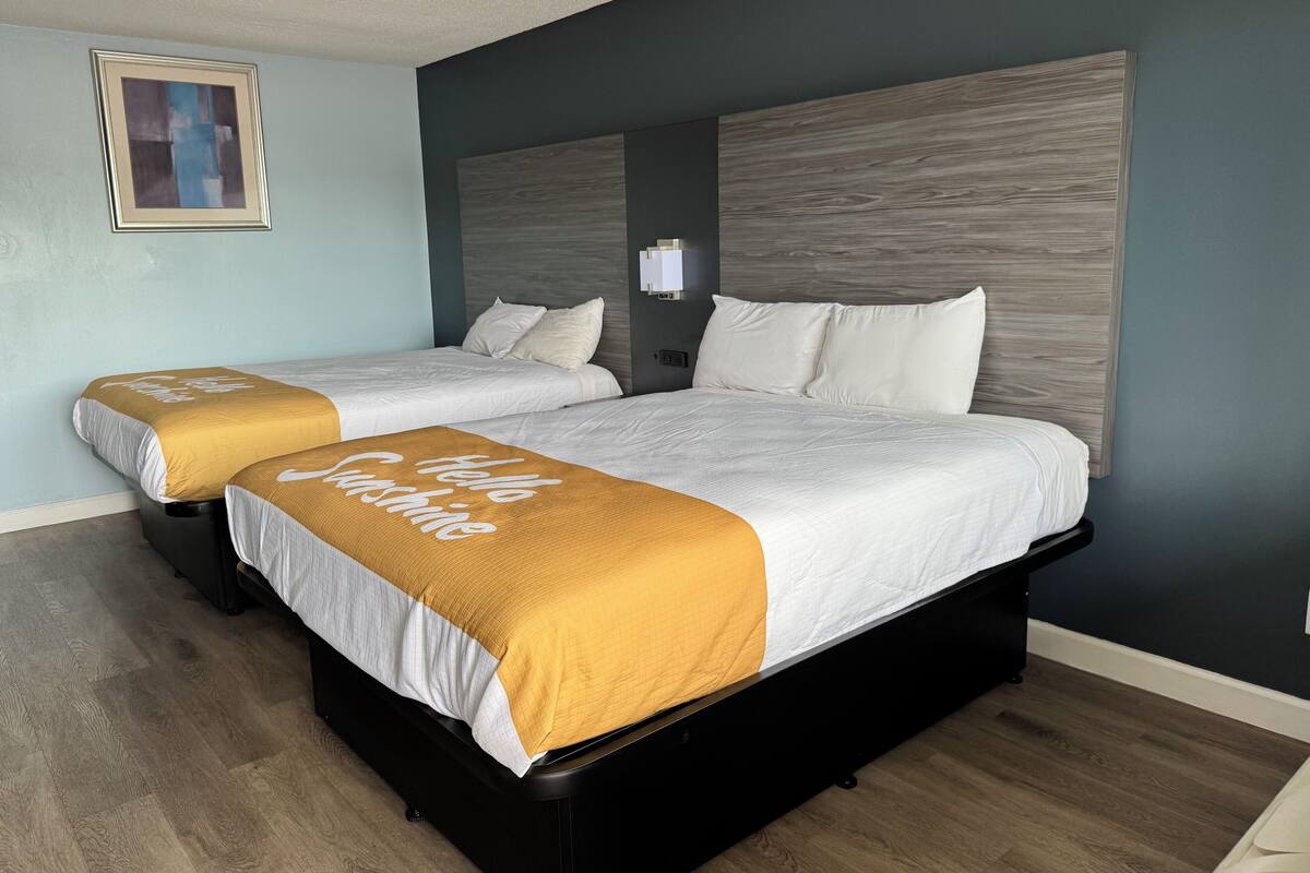 A hotel room with two beds, each with a white and mustard-colored bedspread, a dark headboard, and a framed picture on the wall.