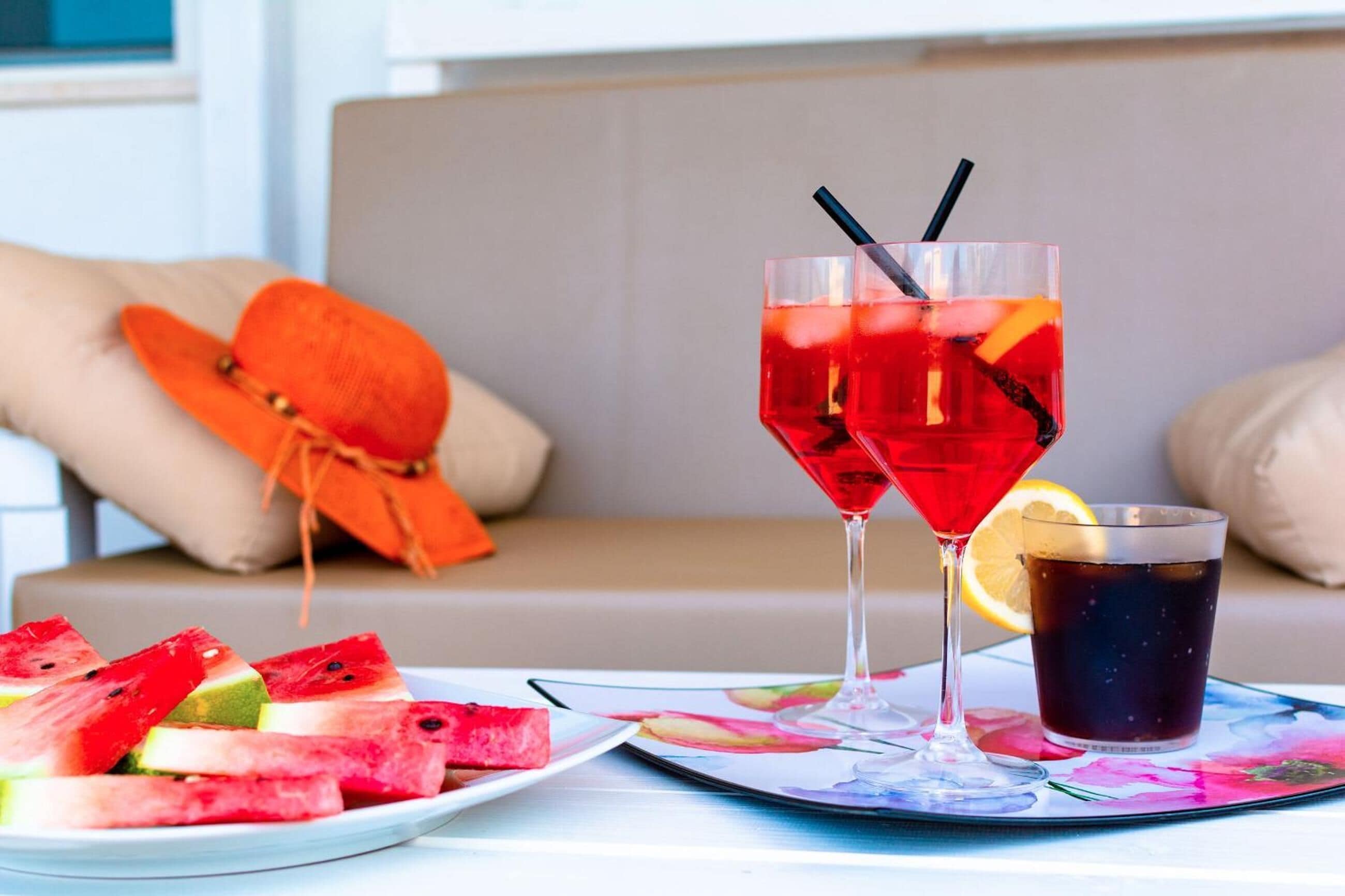A table with two glasses of red drink, a glass of dark beverage, and a plate of watermelon slices.