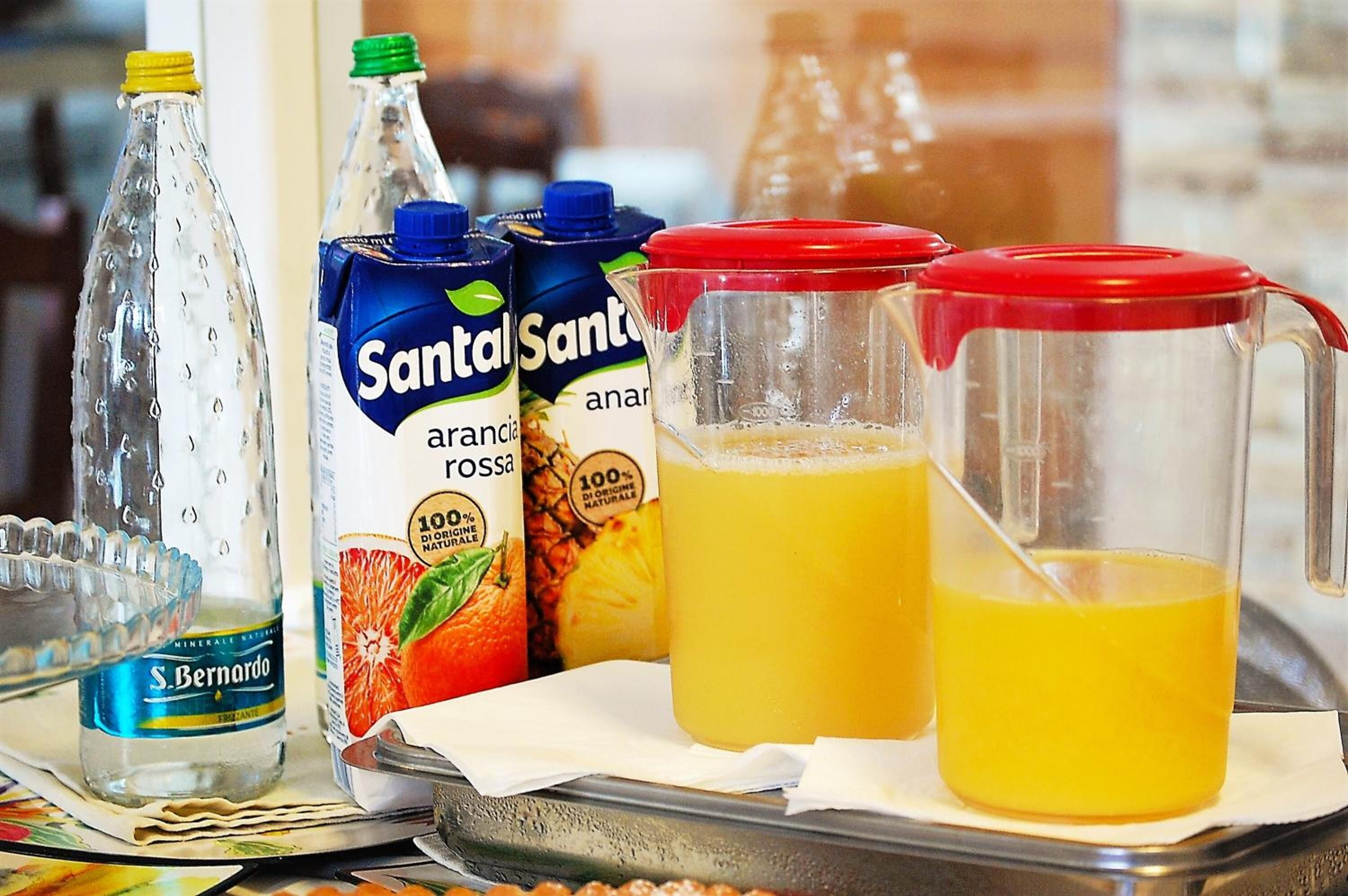 Una bandeja con diversas bebidas, incluyendo una botella de agua S. Bernardo, dos cartones de jugo de piña Santol Santo y una jarra de jugo de naranja.