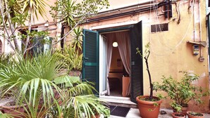 Une cour agrémentée de plantes en pot, une porte avec des volets verts et un bâtiment dont on peut voir les signes d’usure et les canalisations apparentes.