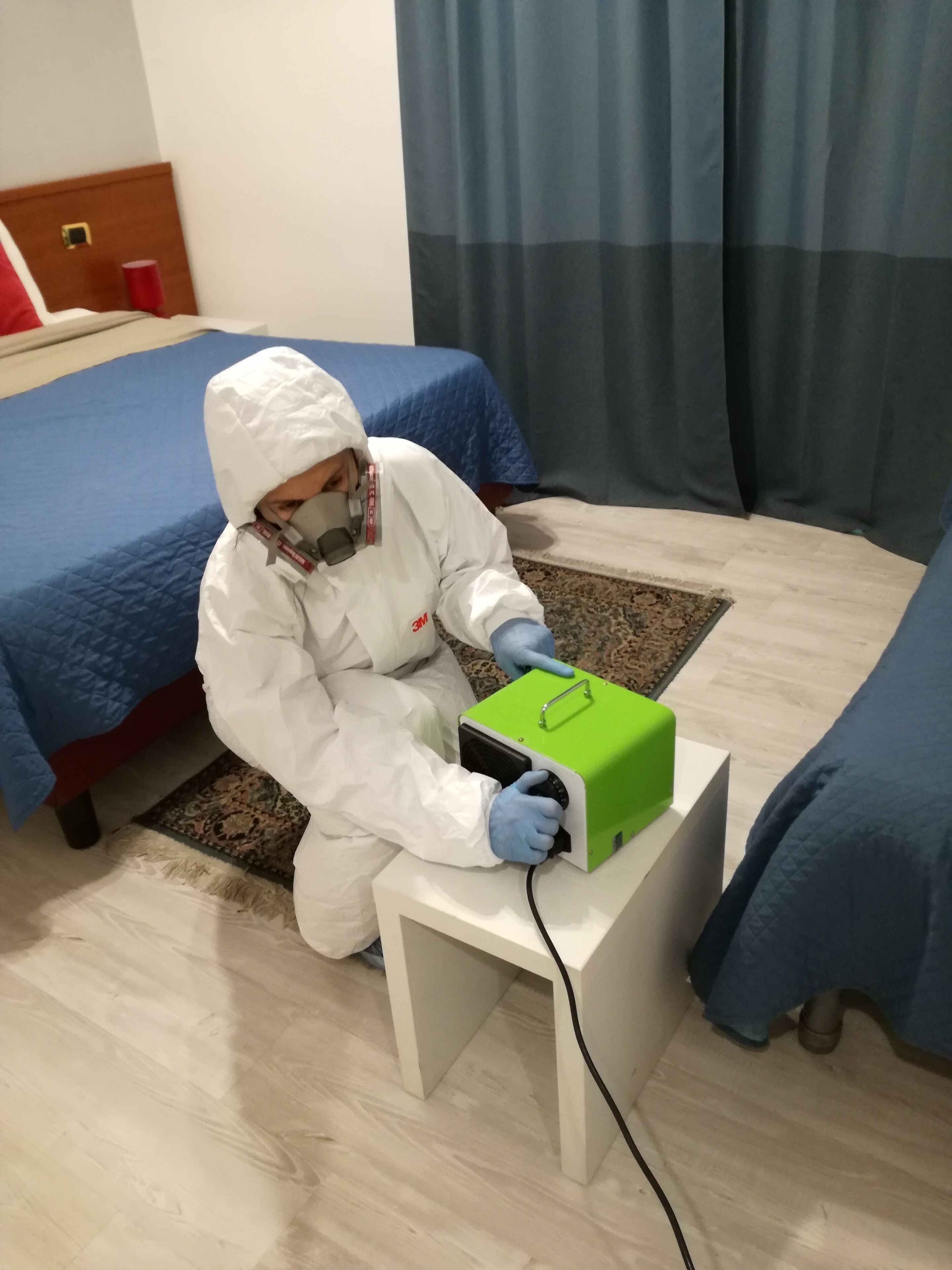 A person in a white protective suit operating a green device on a bedside table in a hotel room.