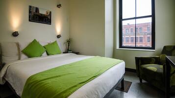 A hotel room with a large bed, green pillows, a nightstand with a lamp, a chair, and a window with a view of buildings.