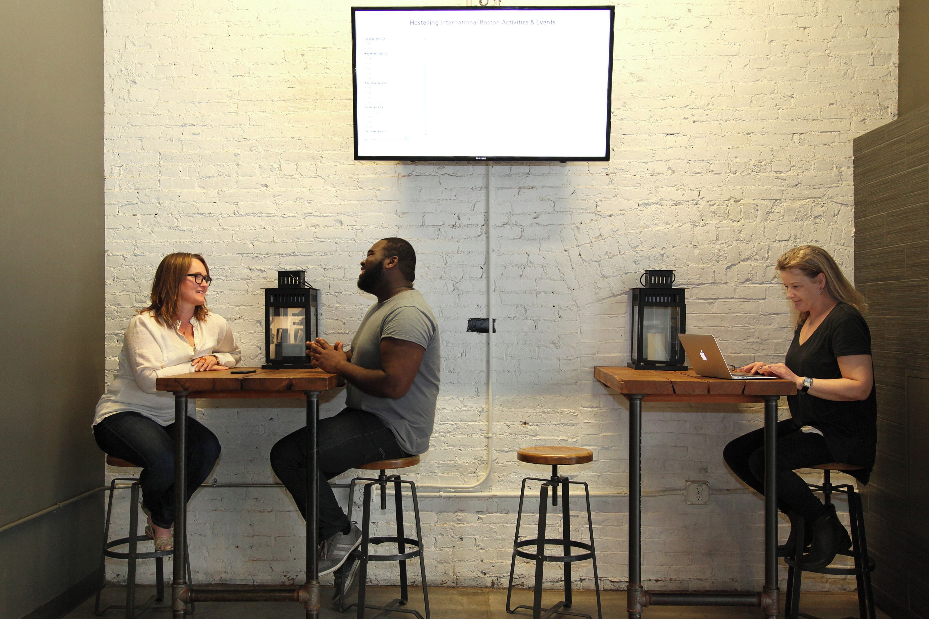 Three people seated at tables with laptops, a projector screen above, and a brick wall in the background.